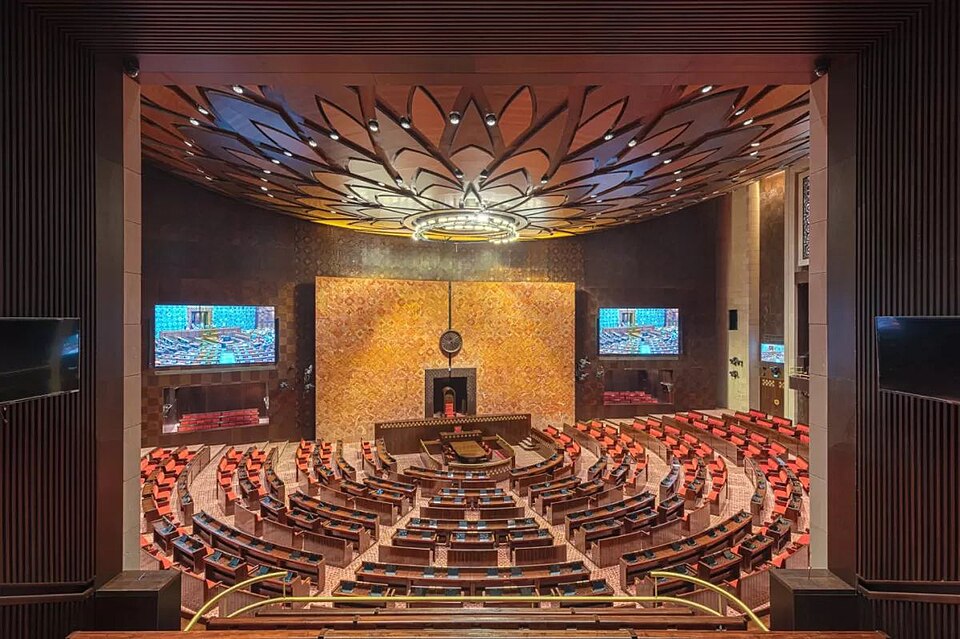 The empty Rajya Sabha chamber in Indias new Parliament building, with curved tiers of red-upholstered members desks facing the Chairmans seat beneath a carved lotus-motif ceiling