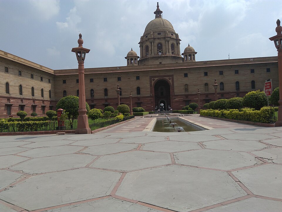 A domed sandstone block of the Secretariat Building on Raisina Hill, New Delhi, seen across a wide paved forecourt with a narrow water channel, clipped hedges and red sandstone lamp columns