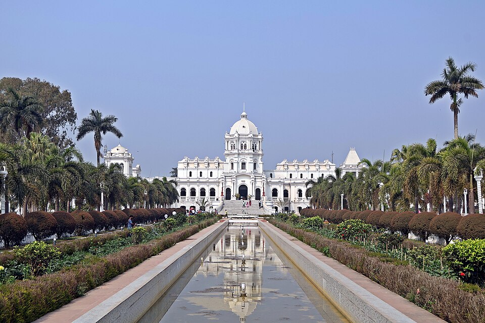 The white domed facade of Ujjayanta Palace in Agartala, Tripura, seen down a long reflecting water channel flanked by palm trees and clipped garden beds