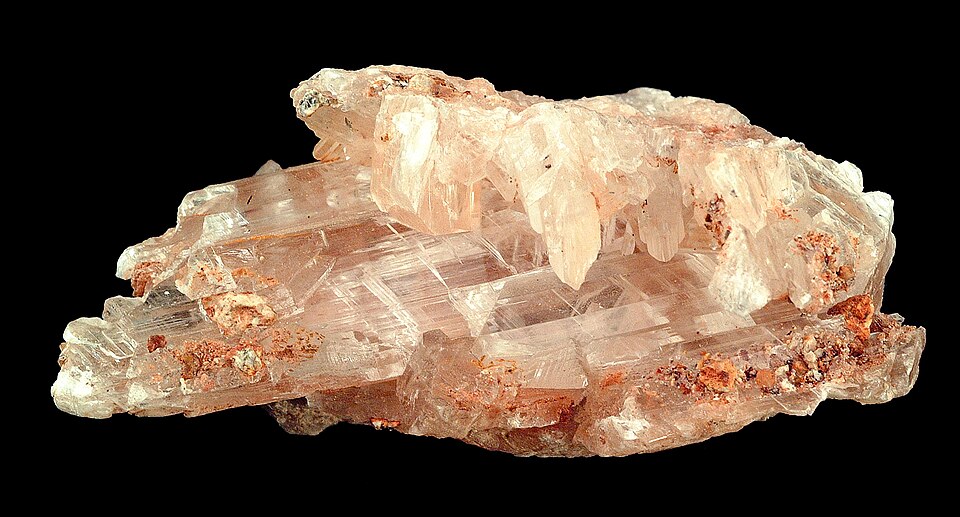 A specimen of gypsum in its selenite form: a cluster of translucent pale-pink bladed crystals with rusty grains of rock clinging to them, photographed against a black background