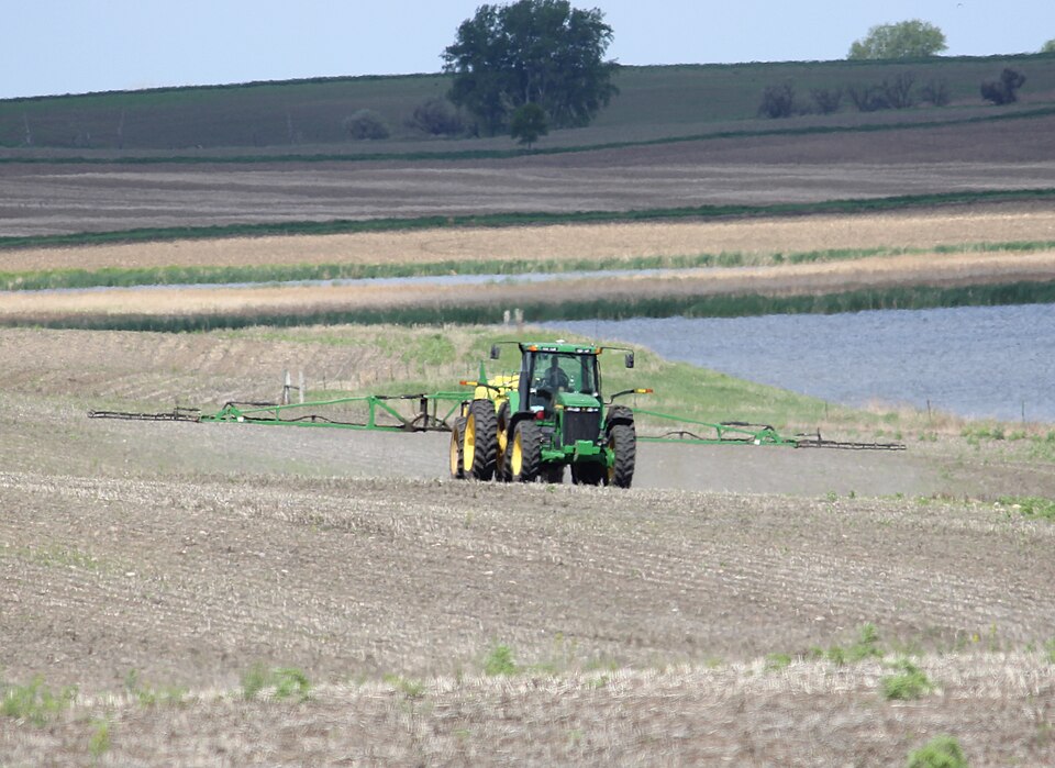 A green tractor towing a wide unfolded spray boom across a farm field, releasing a fine mist of herbicide from nozzles along the boom arms