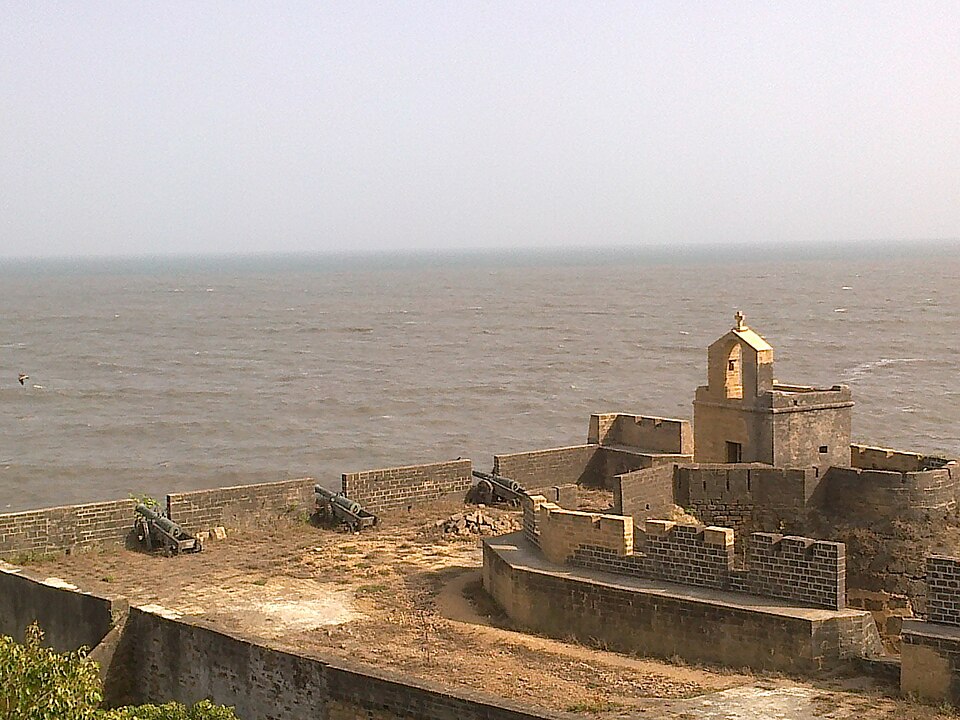The seaward ramparts of Diu Fort: old stone walls with iron cannons mounted along them, a small Portuguese-era bell tower topped by a cross, and the open sea beyond