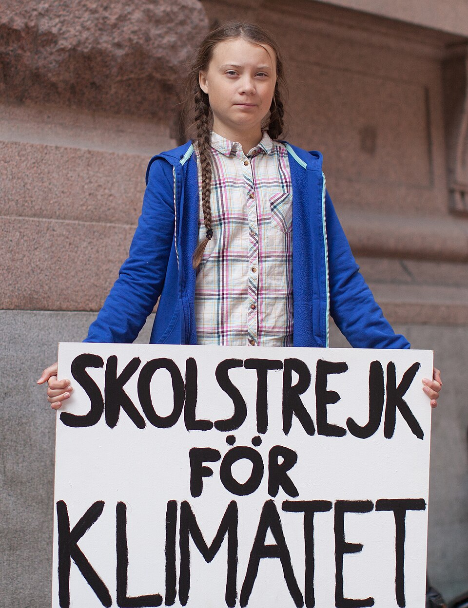 Greta Thunberg, a teenage girl in braided hair and a blue jacket, standing outside a stone building holding a hand-painted placard reading SKOLSTREJK FOR KLIMATET (school strike for the climate)