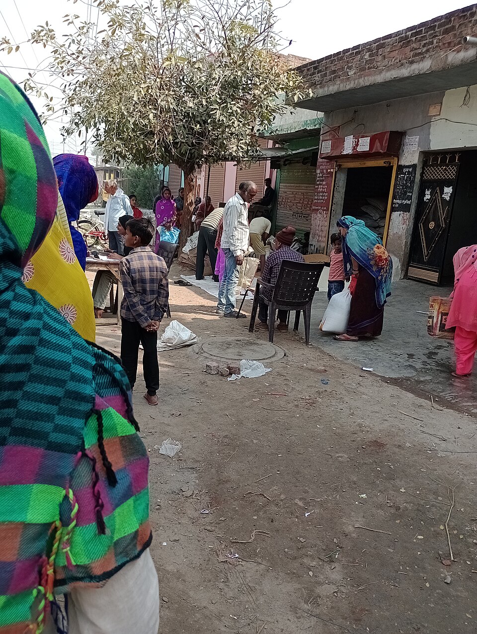 Villagers queuing in a dusty village lane outside a small government ration shop, a woman walking away with a white sack of grain while a clerk sits writing at a table.