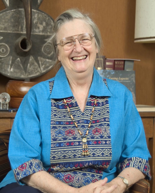 Portrait of Elinor Ostrom, an elderly woman with grey hair and glasses in a blue embroidered tunic, smiling broadly while seated in an office lined with books