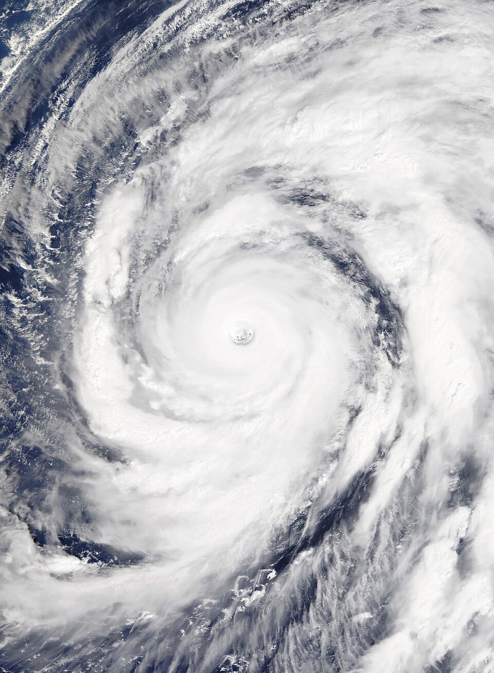A satellite view of a vast white spiral tropical cyclone over dark blue ocean, with a small clear circular eye at its centre and curved rainbands wrapping around it.