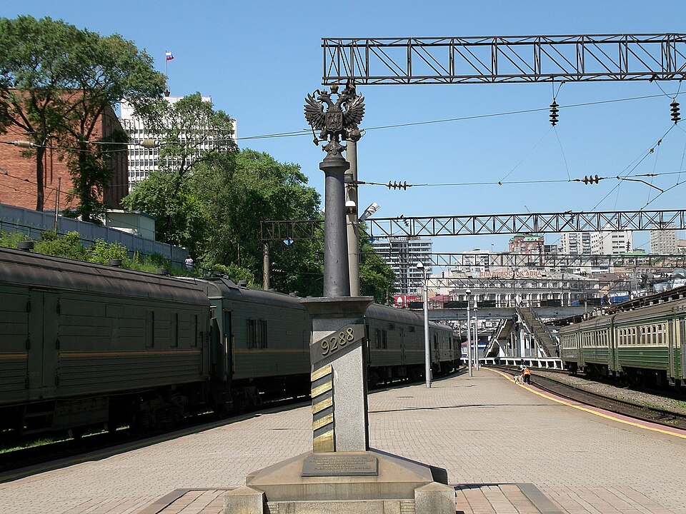 A stone obelisk carved with the number 9288 and topped by a Russian double-headed eagle, standing at the end of a railway platform with green passenger trains on either side.