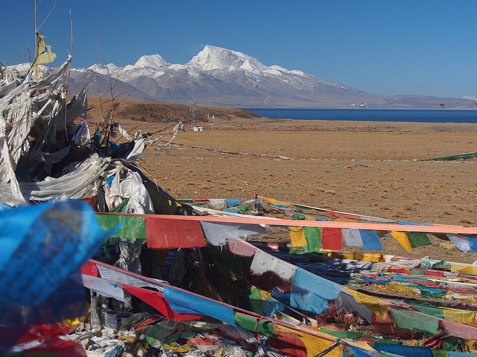 A broad snow-capped Himalayan massif rising beyond the deep blue water of a high-altitude lake, with strings of coloured Tibetan prayer flags strung across the brown plain in the foreground.