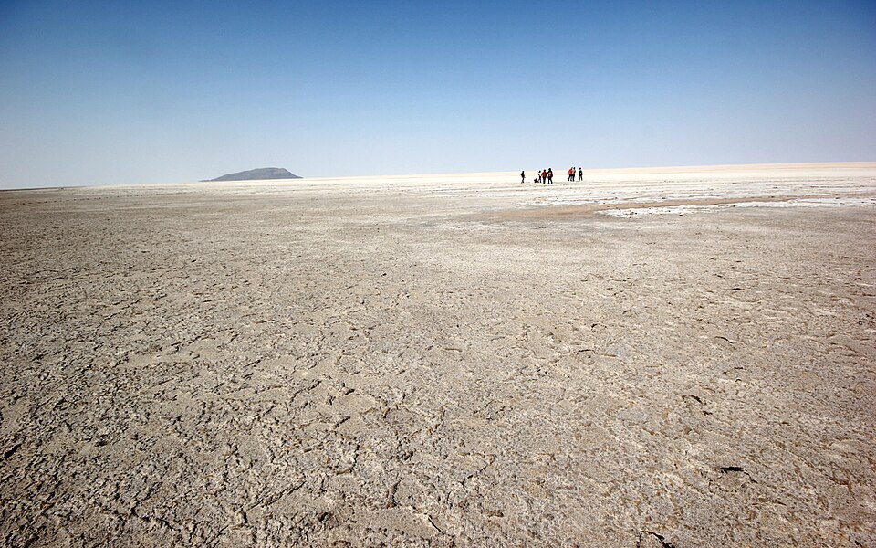 A vast flat expanse of cracked white salt crust stretching to the horizon under a clear sky, with a few tiny distant figures and a low dark hill far away.
