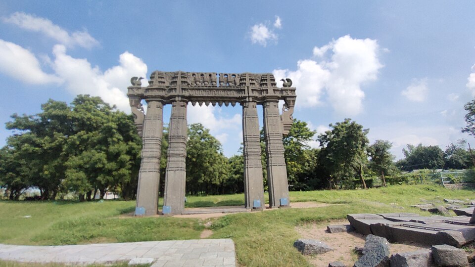 A free-standing carved stone gateway of four thick pillars carrying an ornamented lintel, standing alone on a grassy field beside scattered ruined stone blocks under a blue sky.