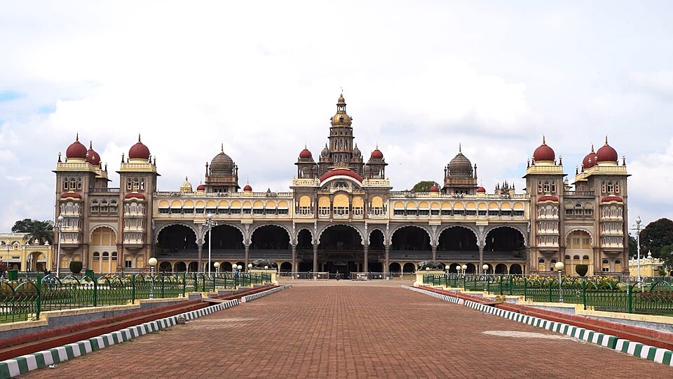 The front facade of Mysore Palace, a long three-storeyed Indo-Saracenic building with grey arcaded verandahs, pink domes and a tall central tower, seen across its brick-paved forecourt