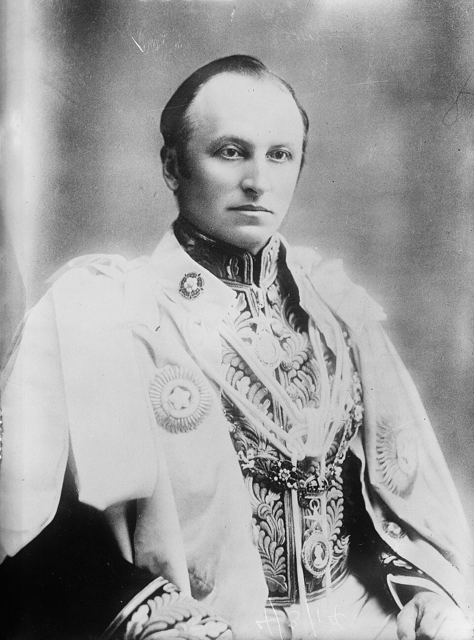A black-and-white studio portrait of a clean-shaven man with hair swept back, wearing an elaborately embroidered court uniform under a heavy ceremonial mantle marked with the star of an order of chivalry.