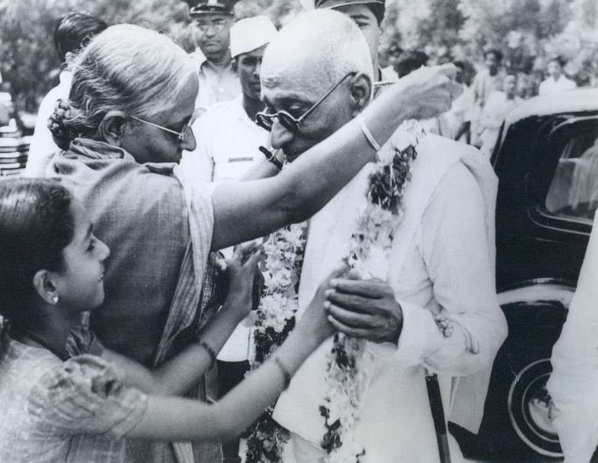 A black-and-white press photograph of an elderly bald man in dark round spectacles and white khadi being garlanded with flowers by a woman as a girl looks on beside a car.