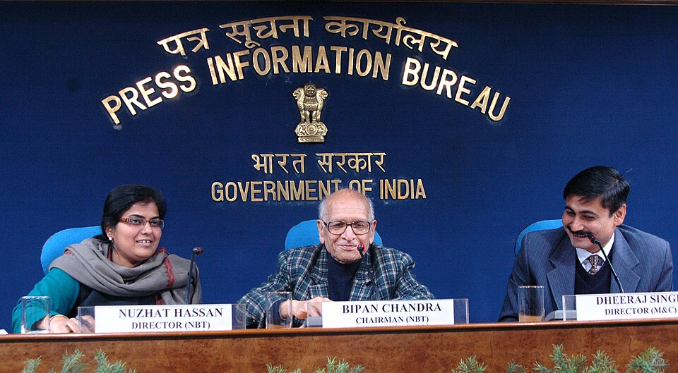 An elderly man in glasses and a checked jacket seated at a Press Information Bureau press-conference table behind a nameplate reading Bipan Chandra, Chairman NBT, with two other officials on either side.