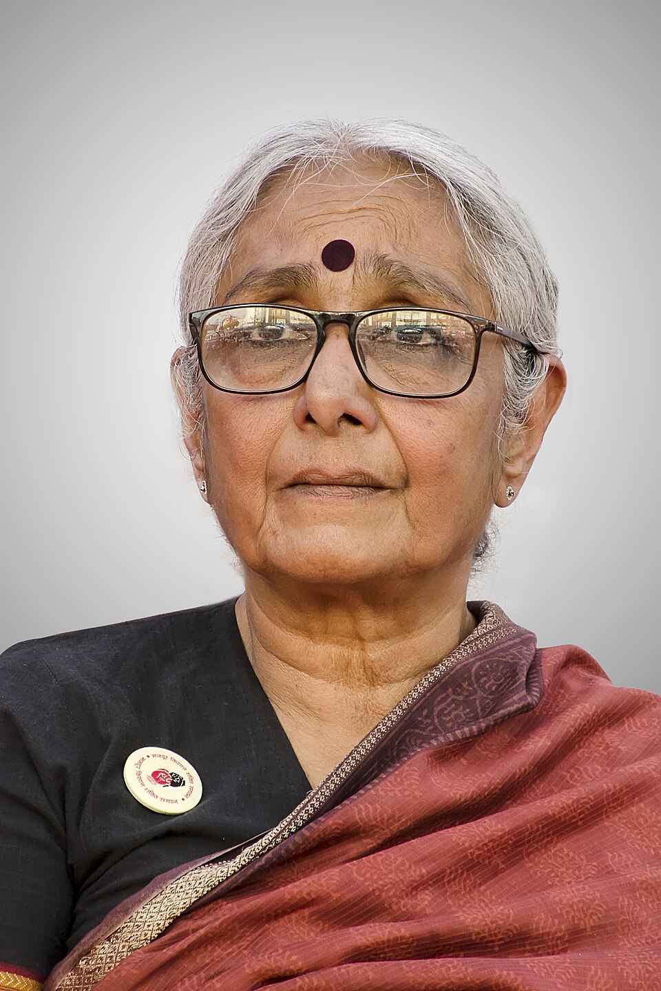 A close portrait of an elderly woman with short white hair, rectangular glasses and a dark red bindi, wearing a black blouse with a round campaign badge and a maroon sari.