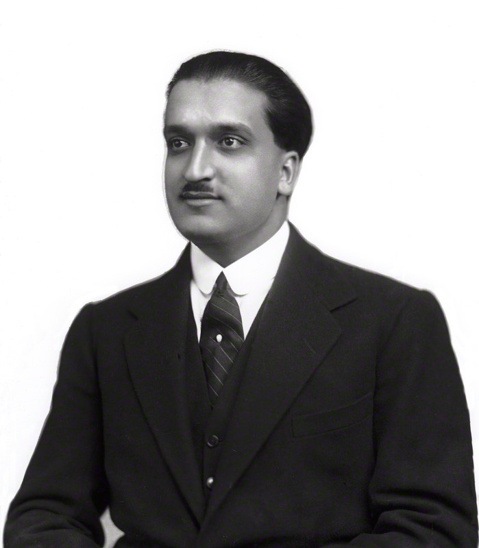A black-and-white studio portrait of a young man with a trimmed moustache and hair combed back, wearing a dark three-piece suit and a striped tie.