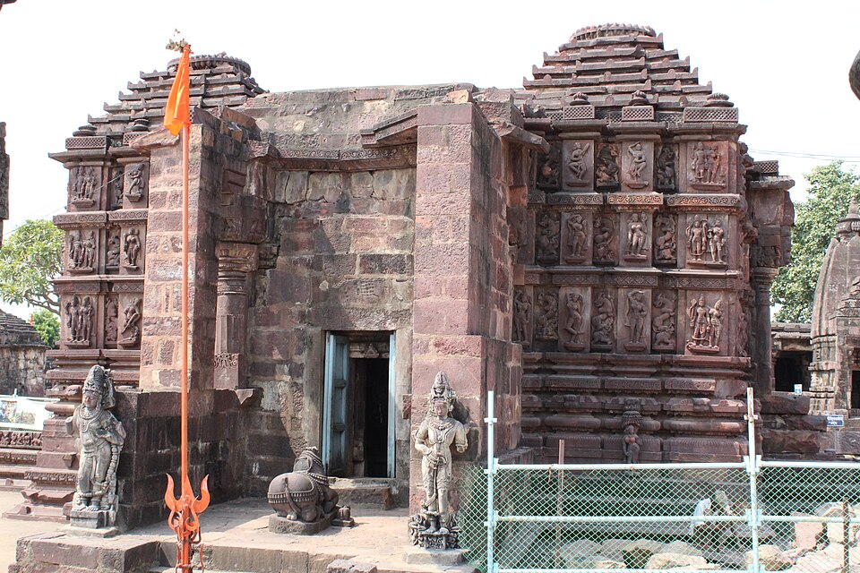 A weathered red sandstone temple with a tiered stepped spire, its outer walls covered in tiers of carved human figures, a Nandi bull and two guardian statues standing before the dark doorway.