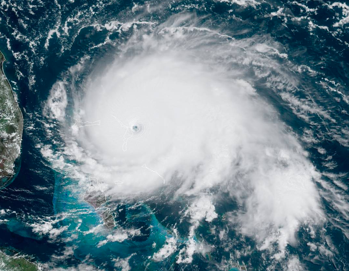 A true-colour satellite view of a large white hurricane with a small clear eye sitting over turquoise island banks, with a green coastline at the left edge of the frame.