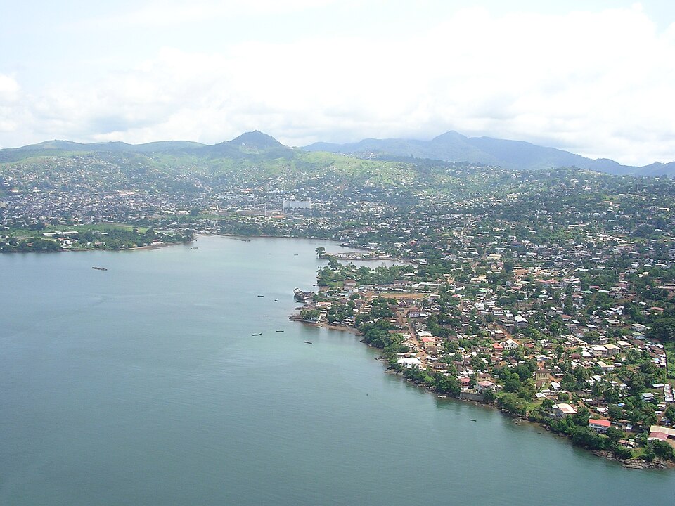 An aerial view of a coastal African city spread along a wide grey-green bay, its low houses climbing green hills that rise behind the shoreline.