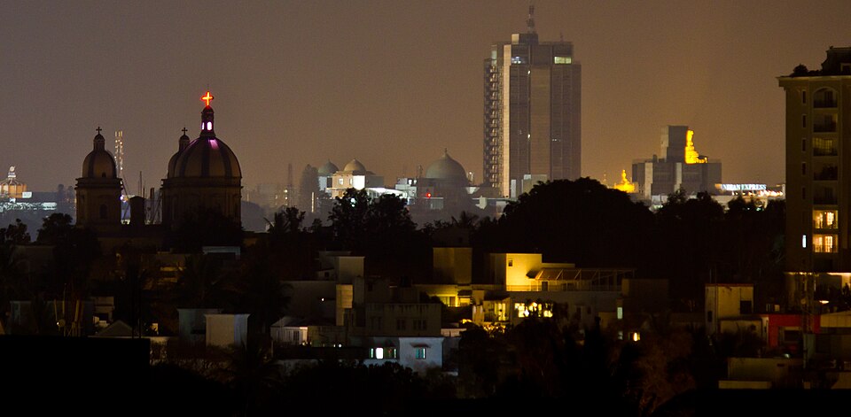 A city skyline at night, with a floodlit church dome topped by a glowing red cross in the foreground and tall office towers and lit low-rise buildings spread behind it.
