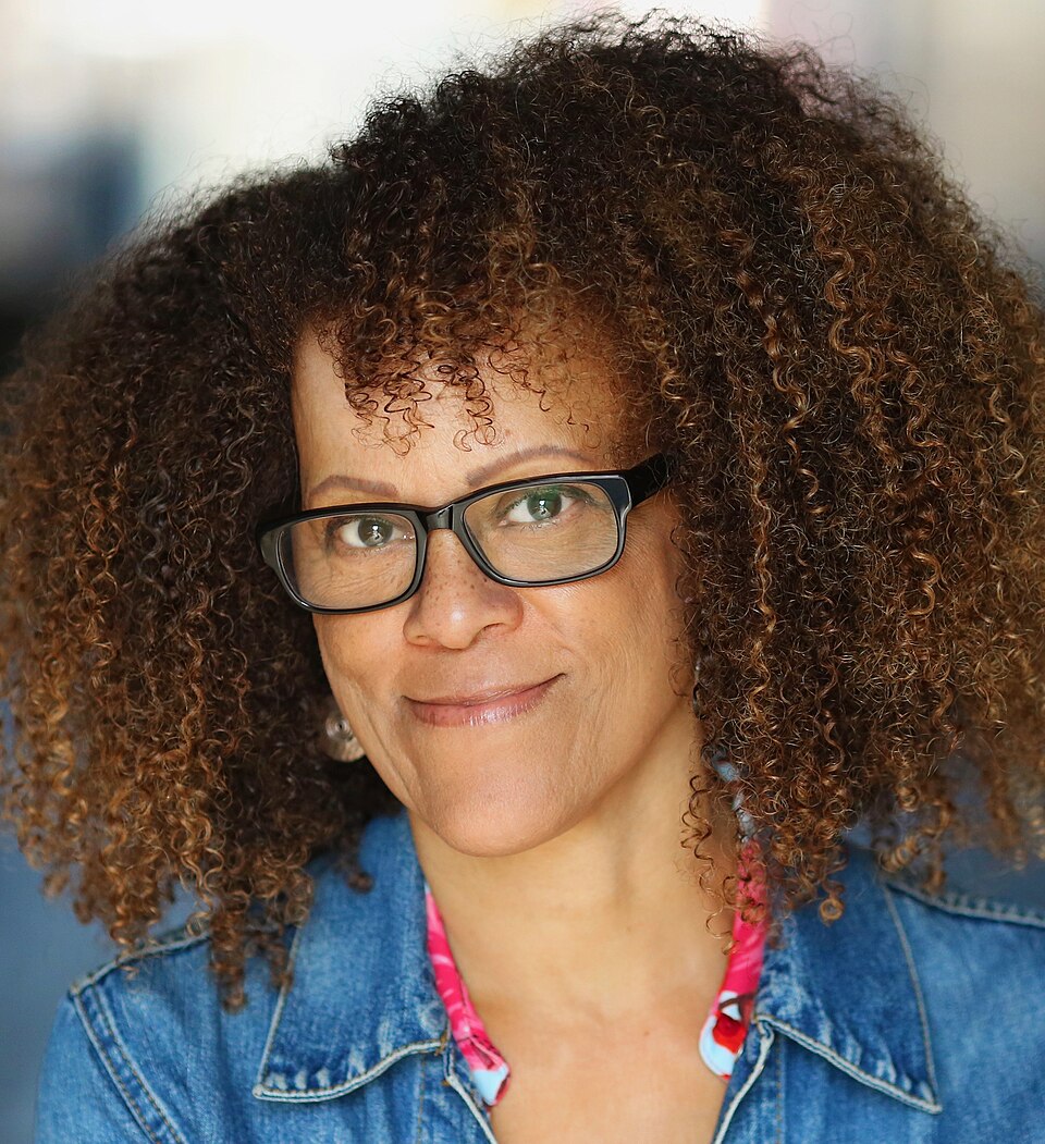 A smiling woman with a large halo of curly brown hair and black-framed rectangular glasses, wearing a denim jacket over a pink patterned top, looking towards the camera.