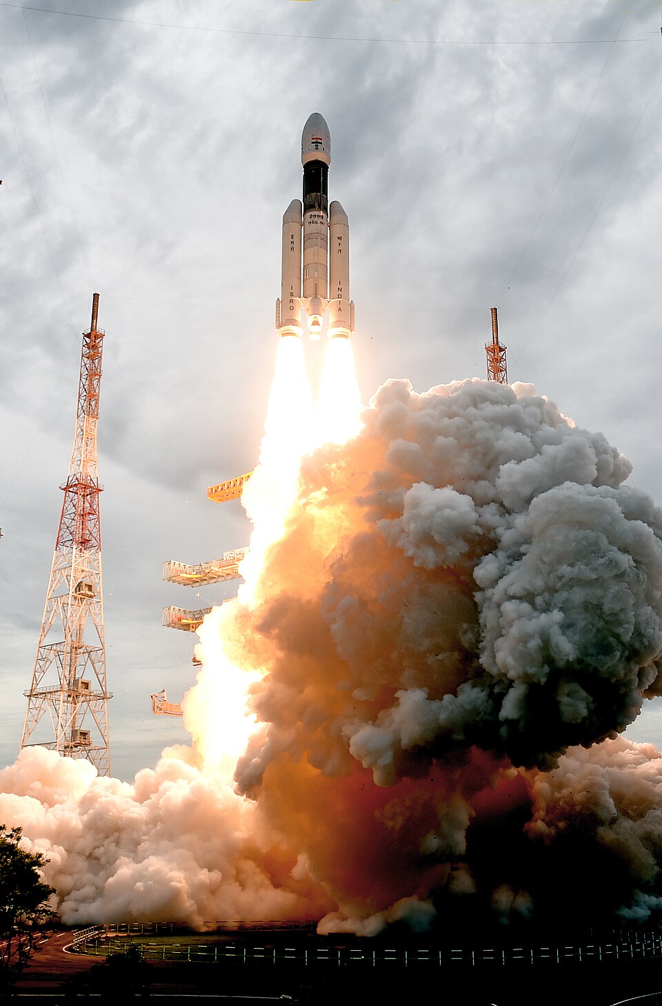 The GSLV Mk III rocket, marked ISRO and INDIA on its two strap-on boosters, climbing off the launch pad on twin plumes of flame amid billowing white exhaust smoke