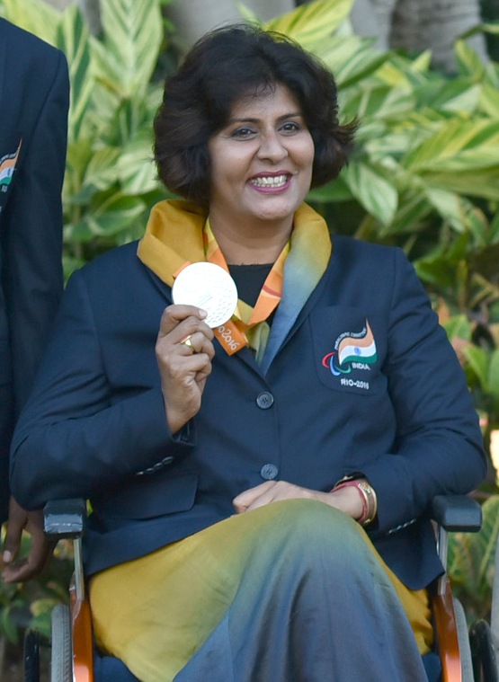 A smiling woman seated in a wheelchair, wearing a navy India team blazer with a Rio 2016 Paralympic crest and a yellow scarf, holding up a silver medal on an orange ribbon.