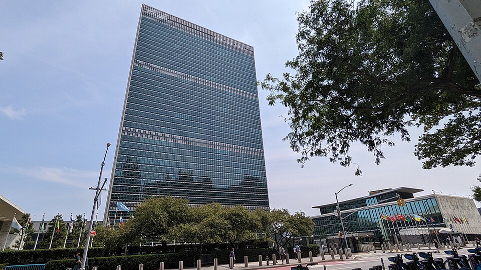 The tall glass-and-steel slab of the United Nations Secretariat Building in New York, with rows of national flags and the low General Assembly and conference wing along the street below.