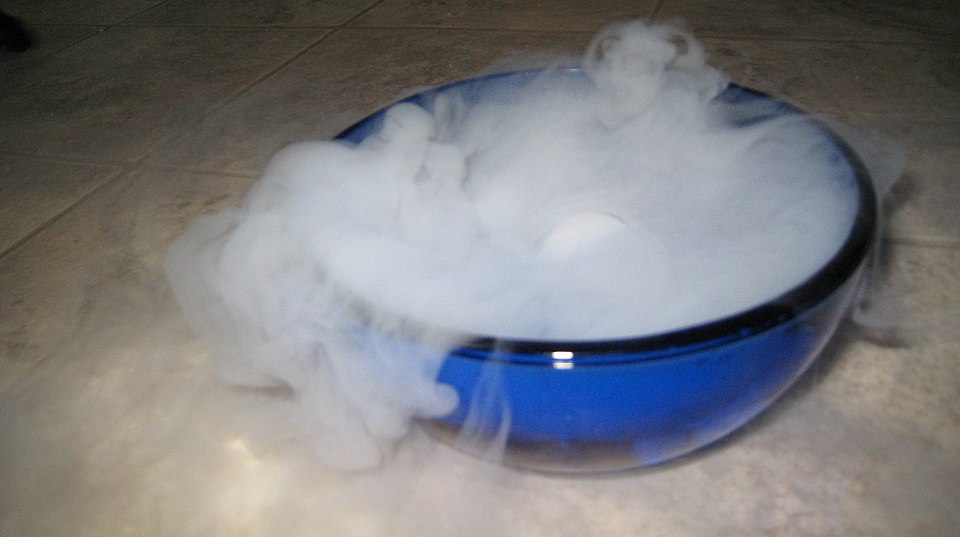 Thick white fog billowing out of a blue glass bowl and spilling over its rim onto a tiled floor, produced by a block of dry ice inside.