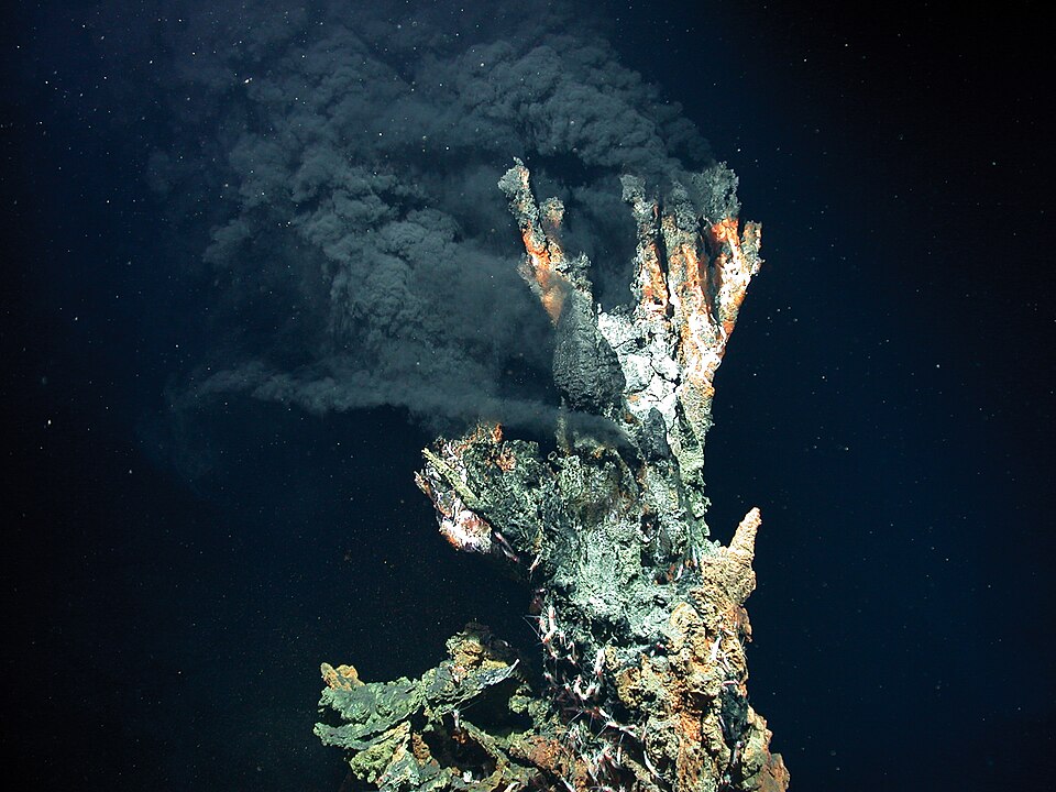 A craggy mineral chimney on the dark deep-sea floor jetting a dense black plume of hot fluid upward into the surrounding water, lit by a submersible lamp.