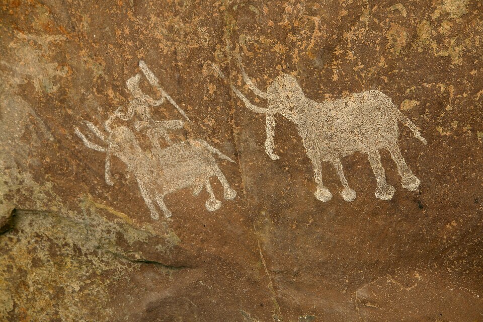 Prehistoric white rock paintings on a brown rock surface depicting a large elephant and a smaller animal ridden by a figure holding a spear