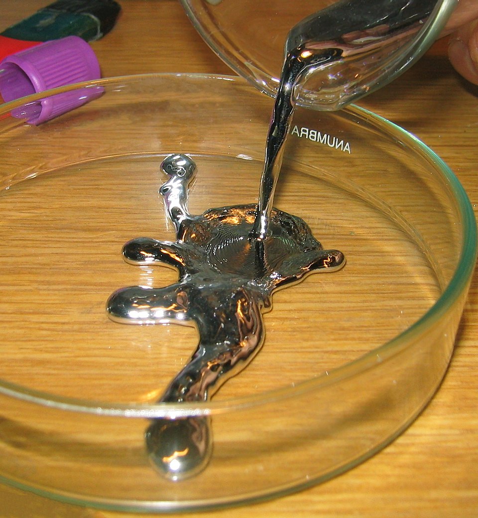 Silvery liquid mercury being poured into a glass petri dish, pooling into shiny metallic globules