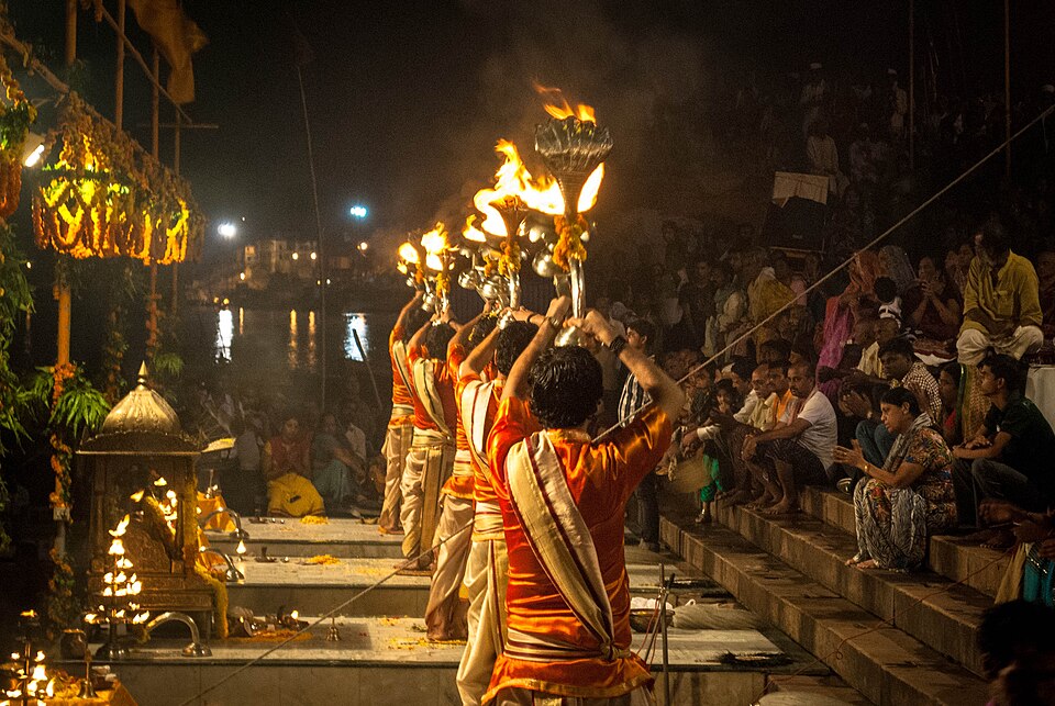 Priests performing the evening Ganga Aarti with tiered flaming lamps at Dashashwamedh Ghat, Varanasi, watched by a seated crowd on the steps