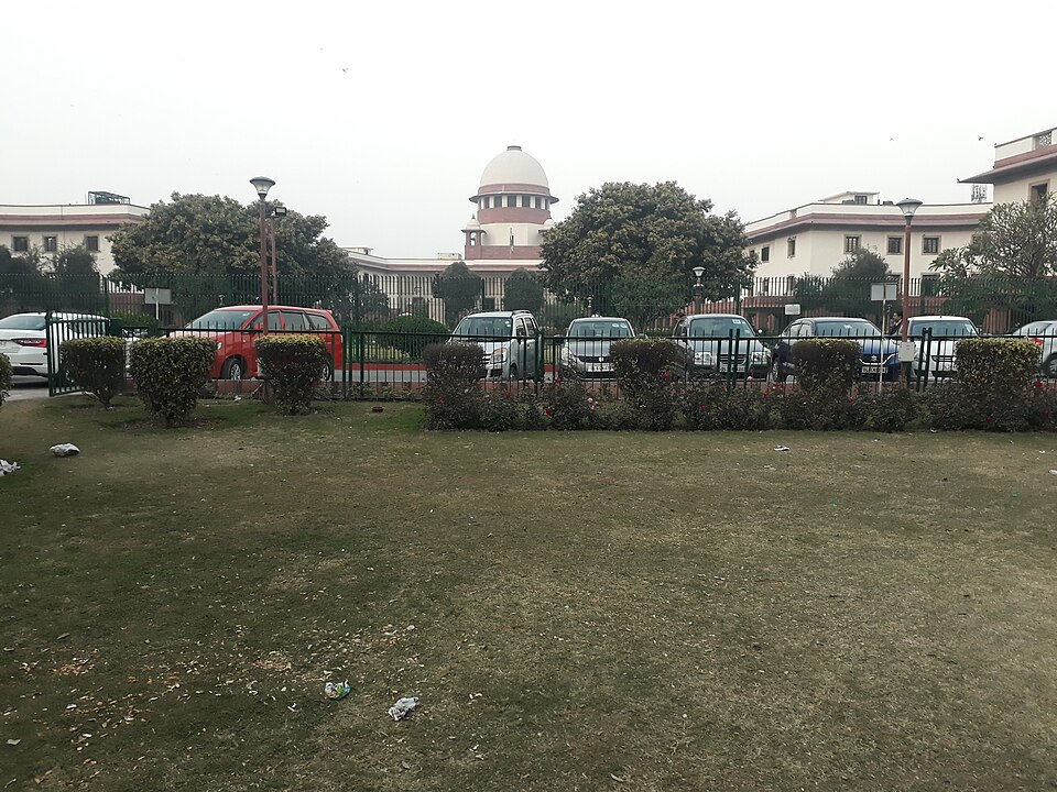 A front view of the Supreme Court of India in New Delhi, its white central dome rising behind the lawn and boundary fence