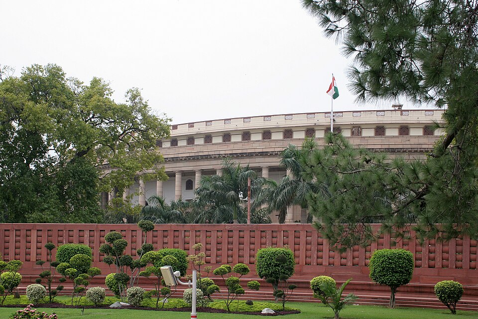 The colonnaded sandstone facade of the circular Sansad Bhavan (Parliament House), New Delhi, with the Indian tricolour flying, behind a red boundary wall and manicured garden