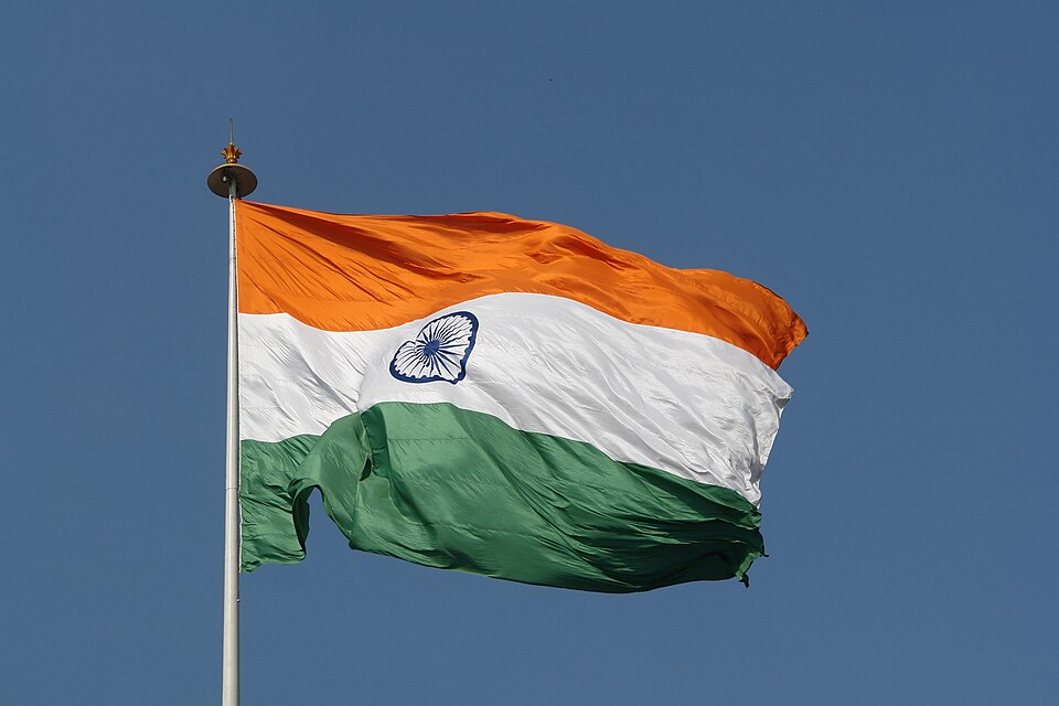 The national flag of India — horizontal saffron, white and green bands with the navy-blue Ashoka Chakra at its centre — flying from a pole against a clear blue sky