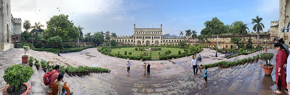 The Bara Imambara complex in Lucknow — its arcaded central edifice rising beyond a walled courtyard garden, with visitors on the surrounding paved terrace