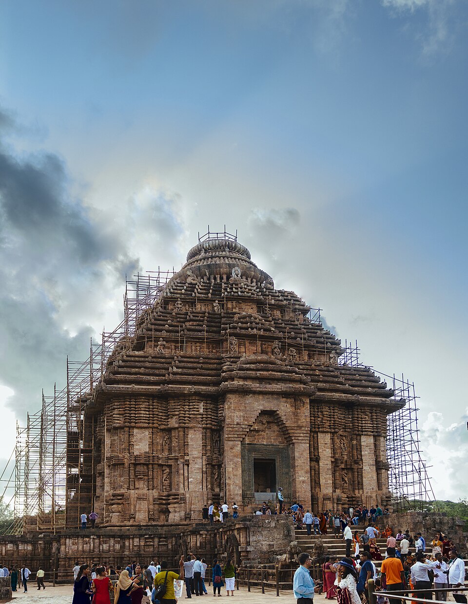 The tiered pyramidal stone jagamohana of the Konark Sun Temple carved with sculptures, with visitors gathered around its base under a cloudy sky