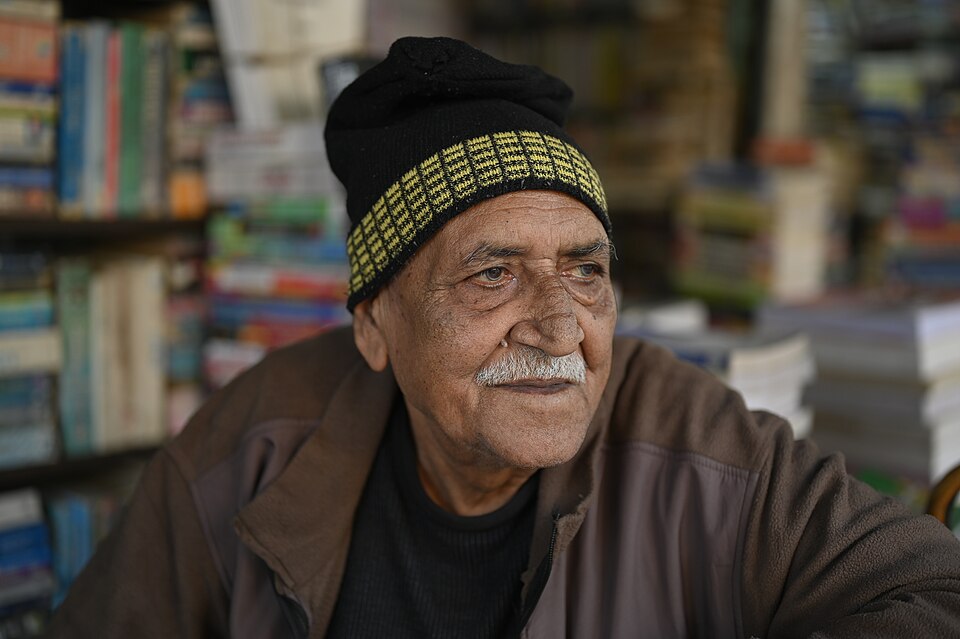 An elderly Indian bookshop owner in a woollen cap seated amid tightly stacked shelves of books in his small retail shop