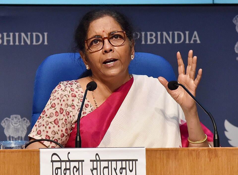 Union Finance Minister Nirmala Sitharaman speaking and gesturing at a government press conference, with her Hindi nameplate on the podium before her