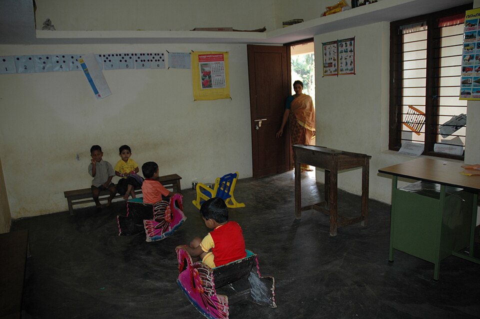 Interior of an anganwadi centre with young children seated on benches and toy rocking horses, numbered and picture charts on the walls, and a worker in a saree at the doorway