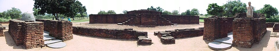 Panoramic view of excavated brick Buddhist monastic ruins with a small stupa on the left and a standing Buddha statue on the right
