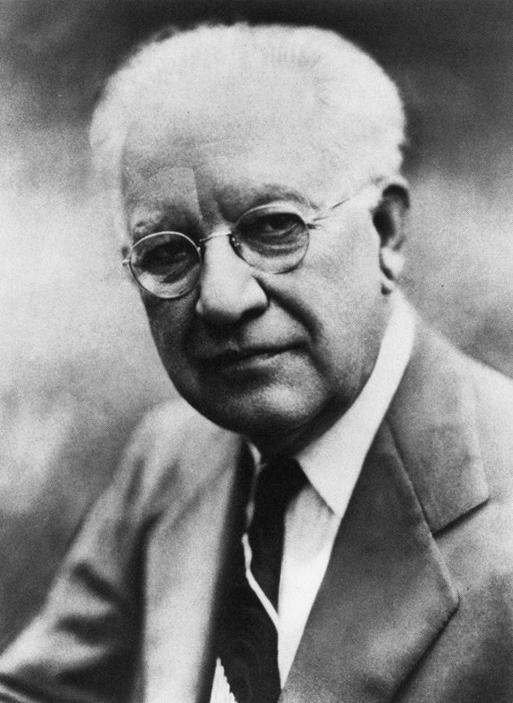 Black-and-white portrait of an elderly white-haired man wearing round spectacles, a suit and tie