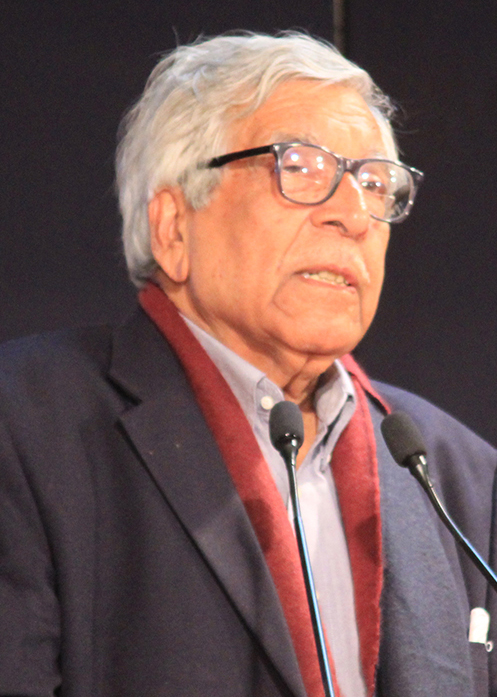 Portrait of an elderly man with white hair and black-framed glasses, wearing a dark blazer and maroon scarf, speaking at a microphone