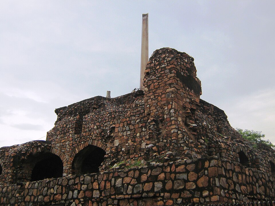 The rubble-stone terraced ruins at Feroz Shah Kotla in Delhi, topped by a polished sandstone Ashokan pillar rising against a grey sky