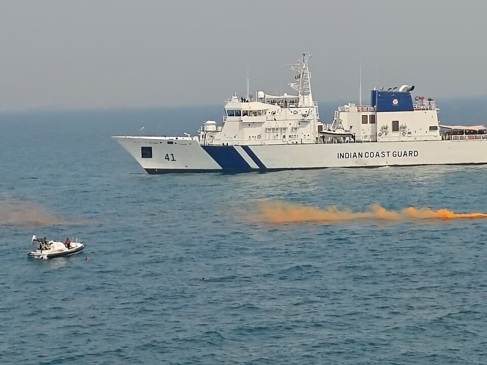 An Indian Coast Guard patrol ship, hull marked INDIAN COAST GUARD, under way at sea with a rigid inflatable boat and an orange smoke marker nearby during an exercise