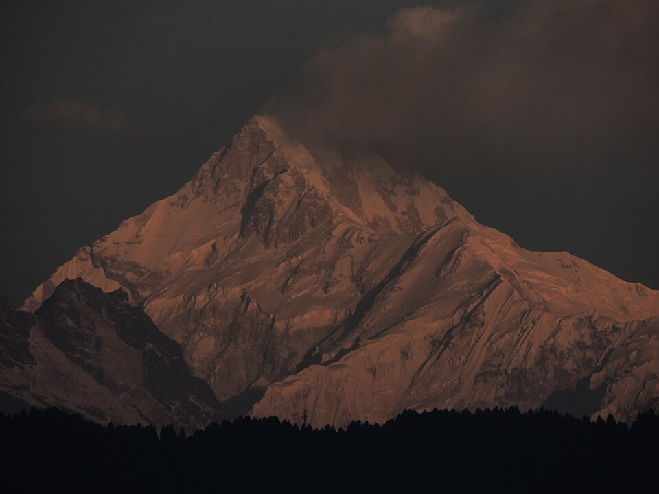 The snow-and-rock summit of Kanchenjunga in the Sikkim Himalaya, lit by warm sunset alpenglow above a dark forested ridge
