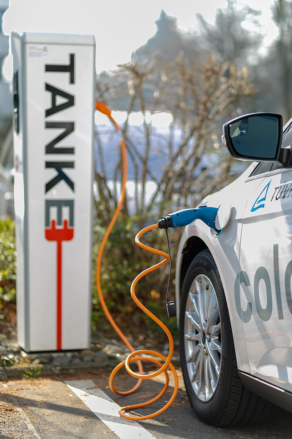 An electric car plugged into a public charging pillar, an orange charging cable running from the station into the car's side charging port
