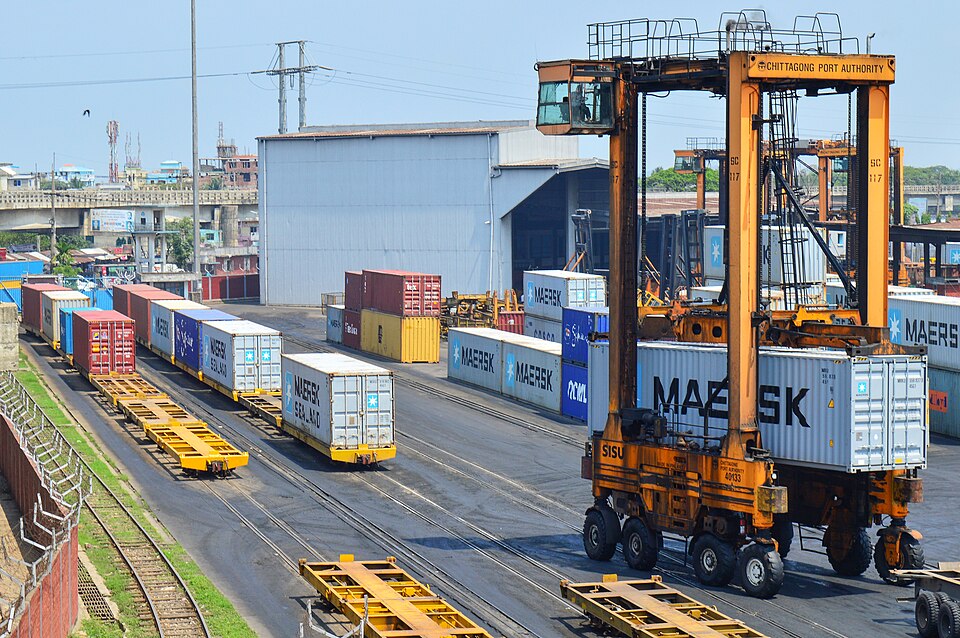 A straddle carrier marked Chittagong Port Authority lifting a shipping container at the Port of Chittagong container terminal, with rail wagons and stacked containers behind