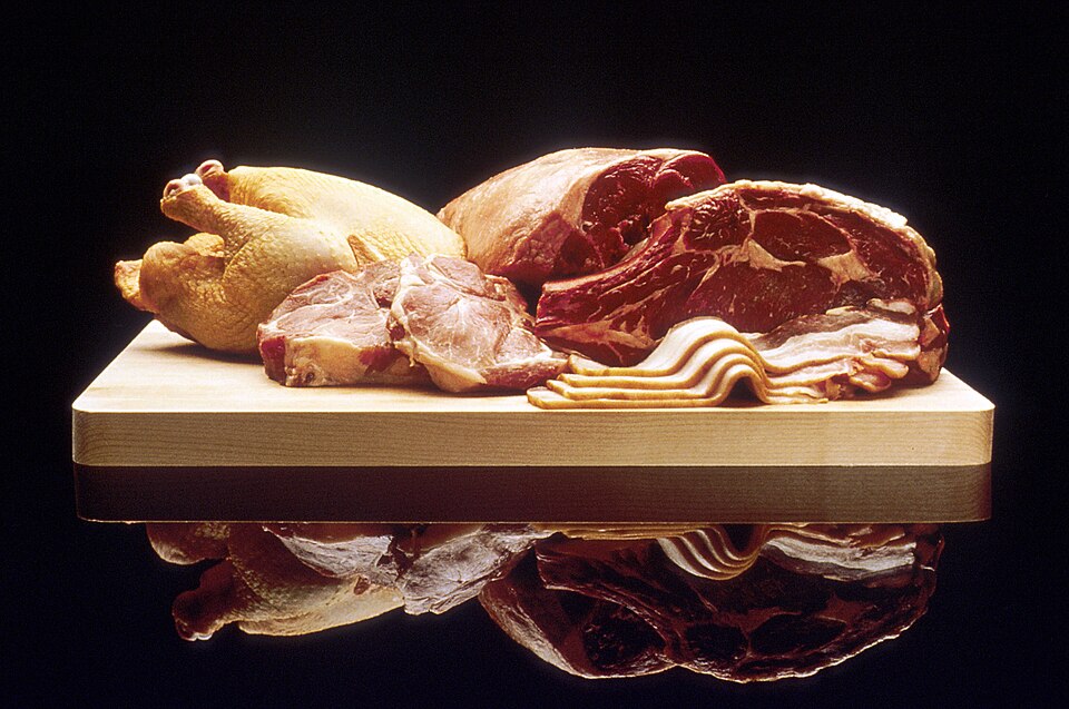 An assortment of raw meats on a wooden board, including a large bone-in beef cut whose deep red colour contrasts with a pale whole chicken